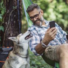 A man enjoying the outdoors with his dog while he uses his mobile phone.