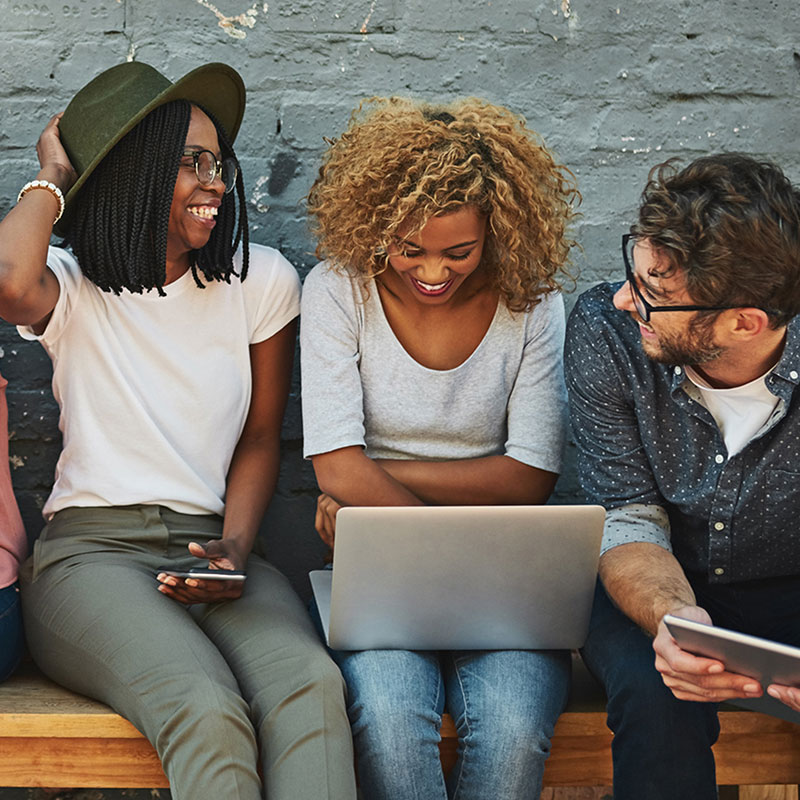 A group of friends laughing while using their mobile phones and laptops.